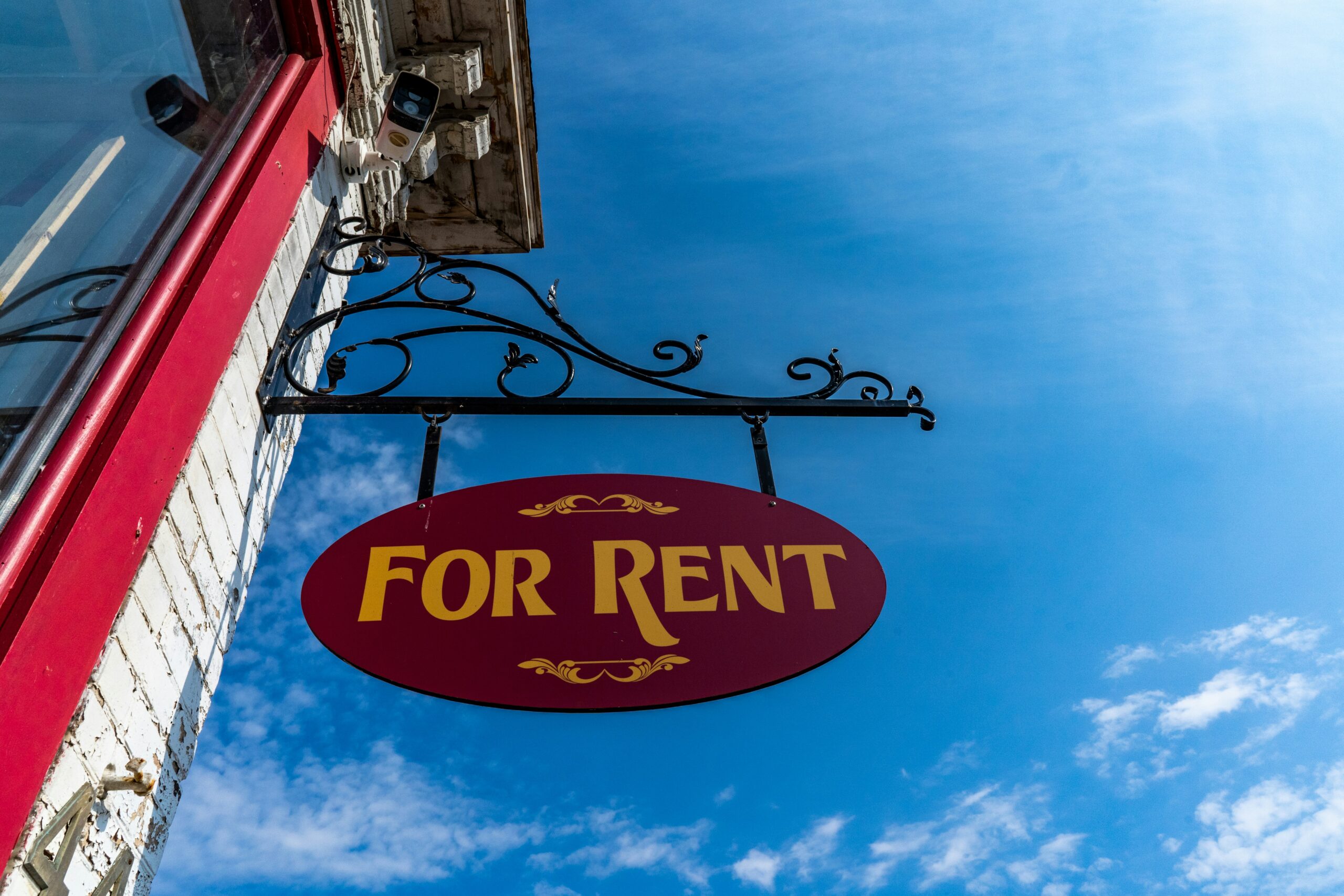 A hanging red and gold “For Rent” sign outside a building under a bright blue sky, representing available small business rental space for lease.