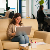 “Employee working on laptop in office lounge while team collaborates in background