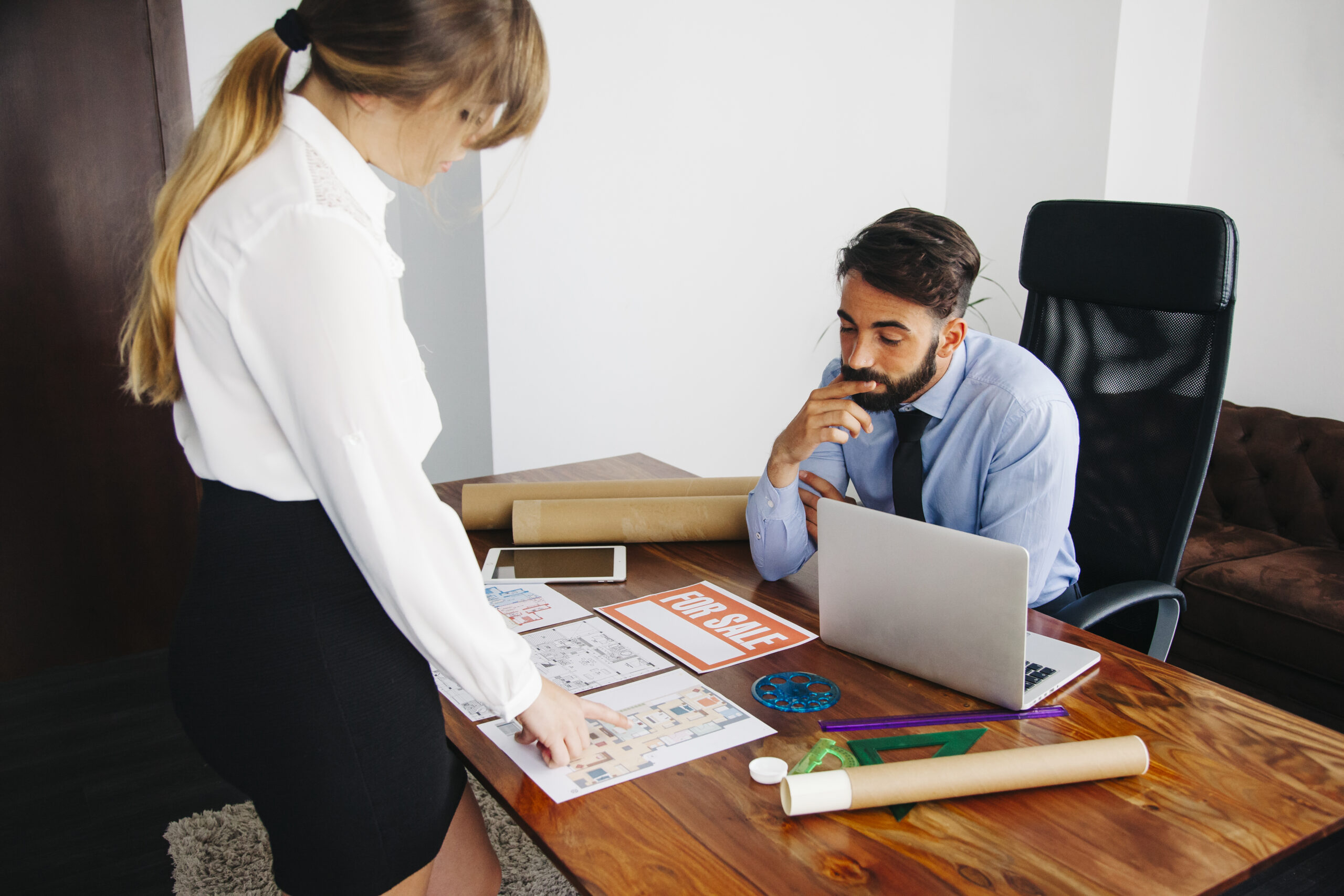 Real estate agent discussing property plans with client, for sale sign on desk