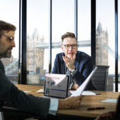 A group of business professionals in a meeting overlooking Tower Bridge in London, with a laptop displaying a "Company's Growth" chart