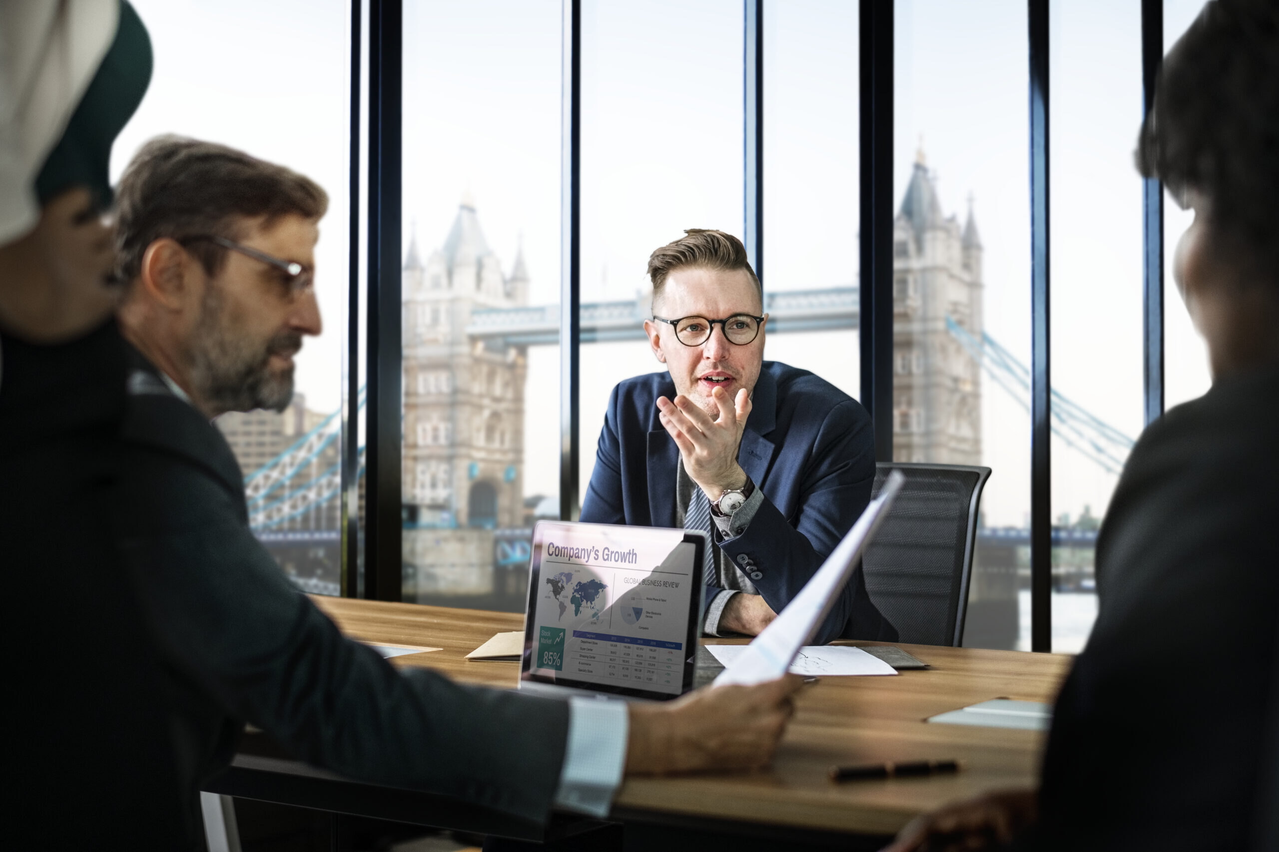 A group of business professionals in a meeting overlooking Tower Bridge in London, with a laptop displaying a "Company's Growth" chart