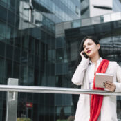 A businesswoman standing outside a modern glass office building, holding a notebook diary while talking on her cellphone.