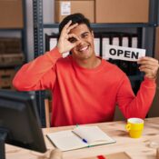 A smiling small business owner holding an open sign at his desk in a rented commercial warehouse space, gesturing an okay sign.