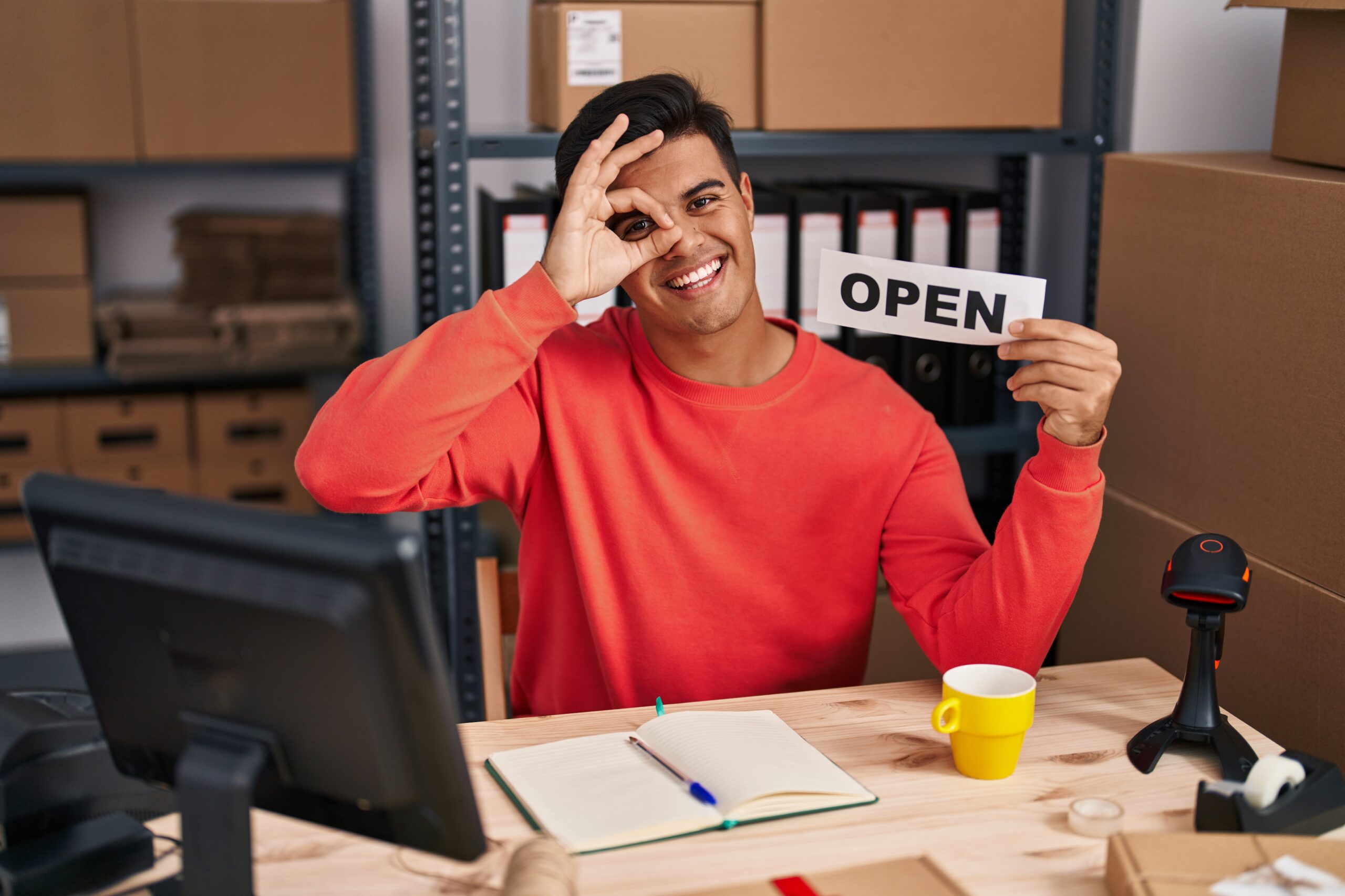 A smiling small business owner holding an open sign at his desk in a rented commercial warehouse space, gesturing an okay sign.