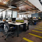 Modern open-plan office with rows of workstations, black mesh chairs, dual monitors, exposed industrial ceiling with pipes and ductwork, glass-walled meeting rooms, and a long corridor with bold yellow striped carpet leading to floor-to-ceiling windows.