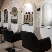 Row of black leather salon styling chairs in front of arched mirrors set into a decorative wallpapered wall, with a wooden staircase and shelving visible in the background of the hair salon.