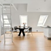 Bright attic-style apartment with sloped ceiling, white ladder leading to a loft space, black round dining table with grey chairs, angled skylight windows, wood flooring, and an open white kitchen area to the right.