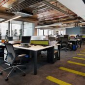 Modern open-plan office with rows of workstations, black mesh chairs, dual monitors, exposed industrial ceiling with pipes and ductwork, glass-walled meeting rooms, and a long corridor with bold yellow striped carpet leading to floor-to-ceiling windows.