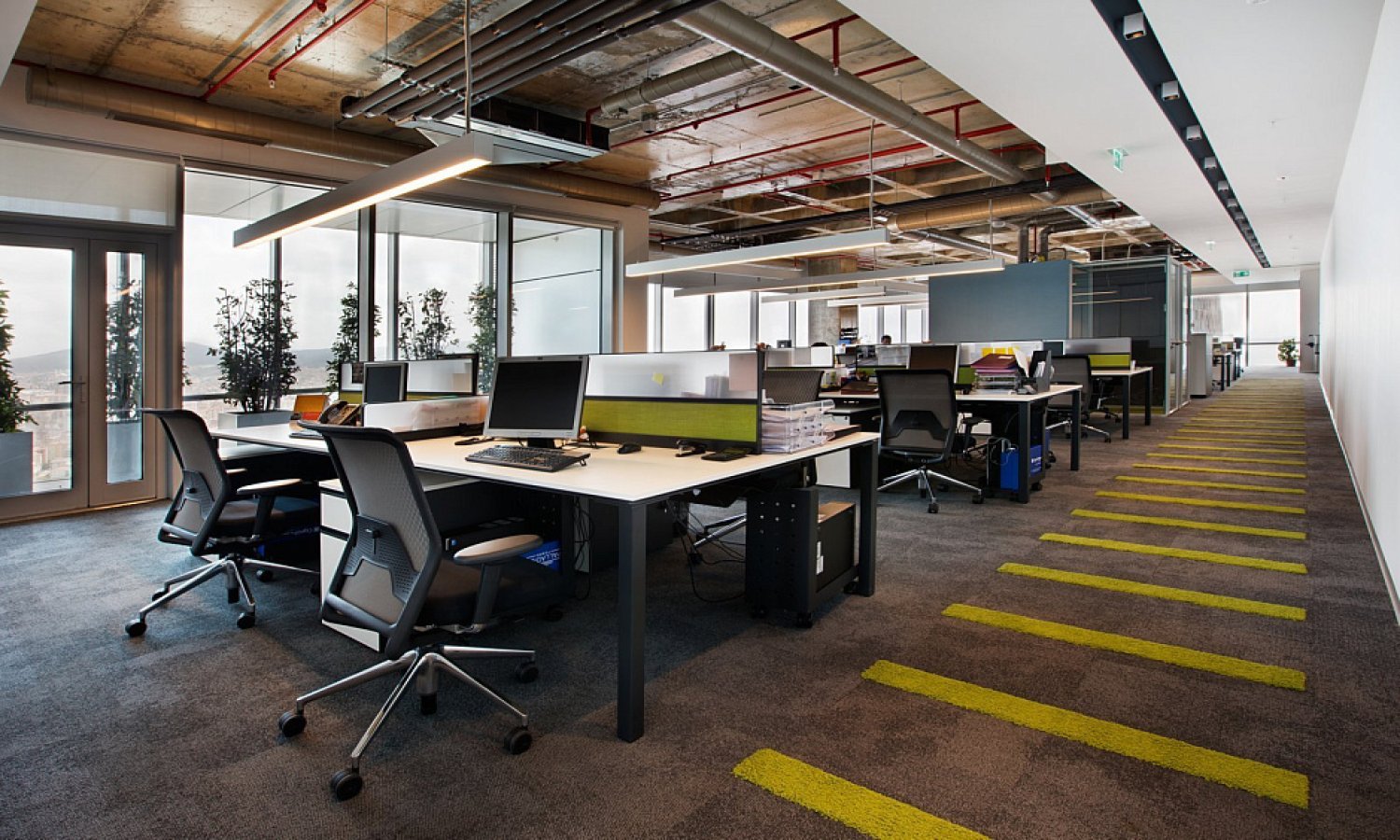 Modern open-plan office with rows of workstations, black mesh chairs, dual monitors, exposed industrial ceiling with pipes and ductwork, glass-walled meeting rooms, and a long corridor with bold yellow striped carpet leading to floor-to-ceiling windows.