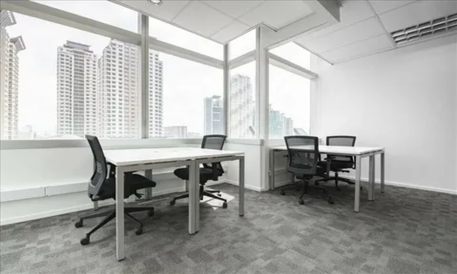 Empty corner office with two white desks and black mesh office chairs, floor-to-ceiling windows offering a city high-rise view, carpeted flooring, and a white ceiling with recessed lighting